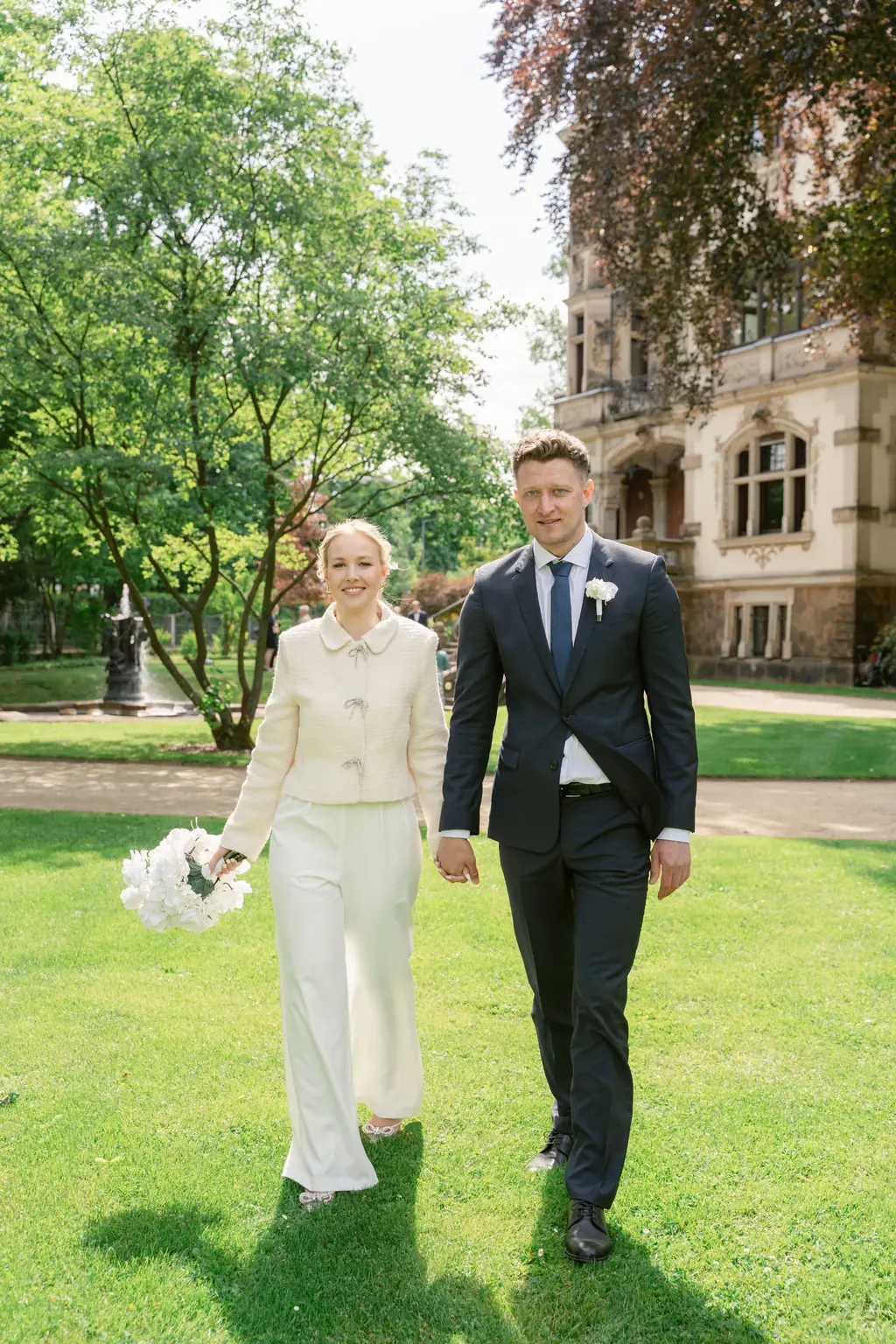 Das Brautpaar spaziert Hand in Hand über die grüne Wiese im Park des Standesamts Goetheallee.