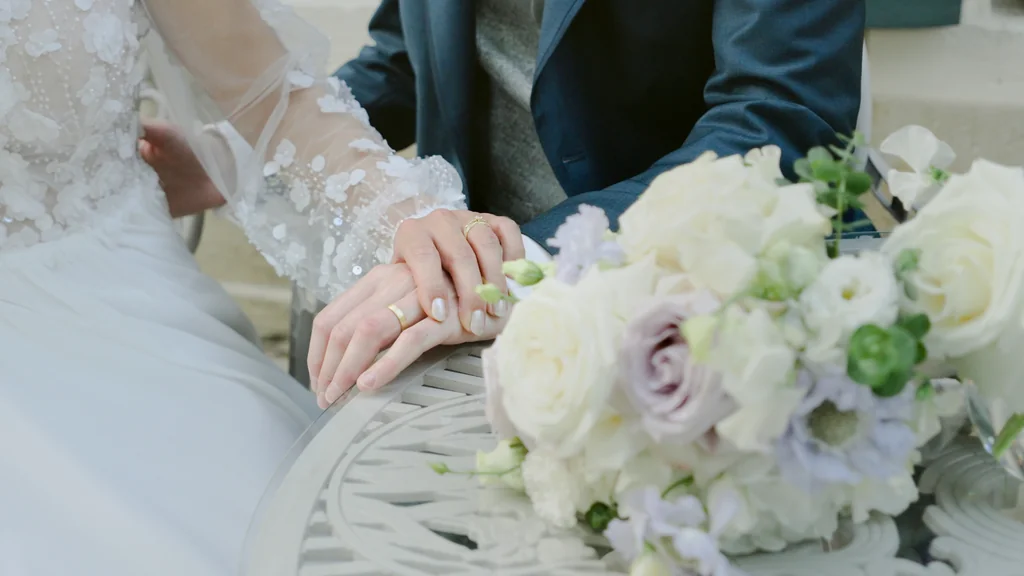 Nahaufnahme der Hände des Brautpaars neben einem hellen Brautstrauß auf einem Eisentisch.