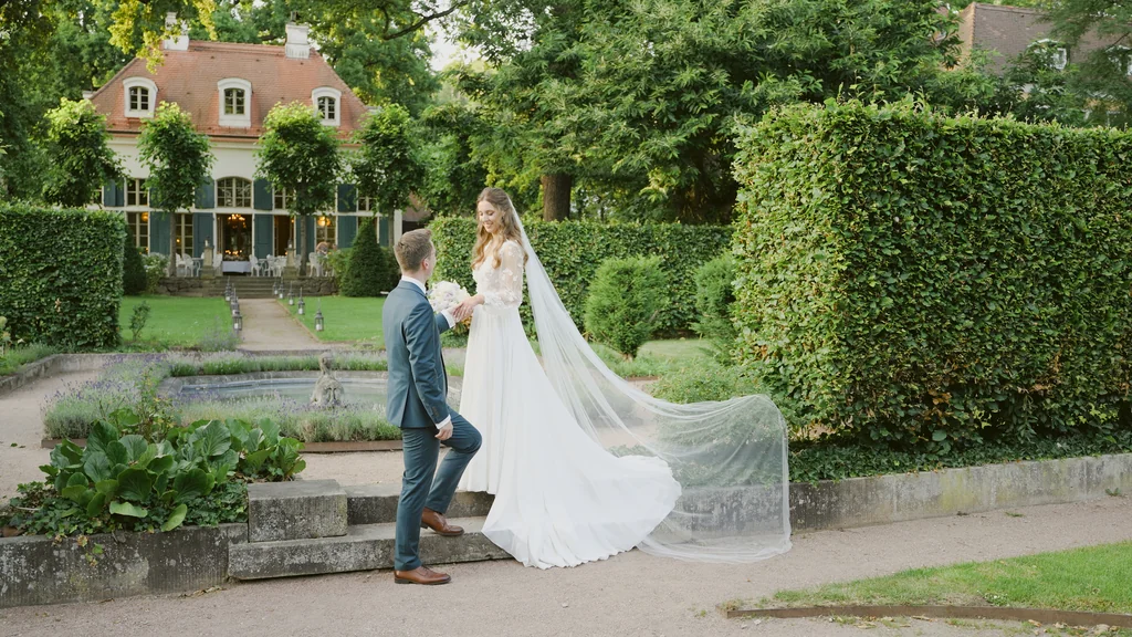 Weitwinkelaufnahme des Brautpaars auf einem Kiesweg im weitläufigen Garten der Villa.