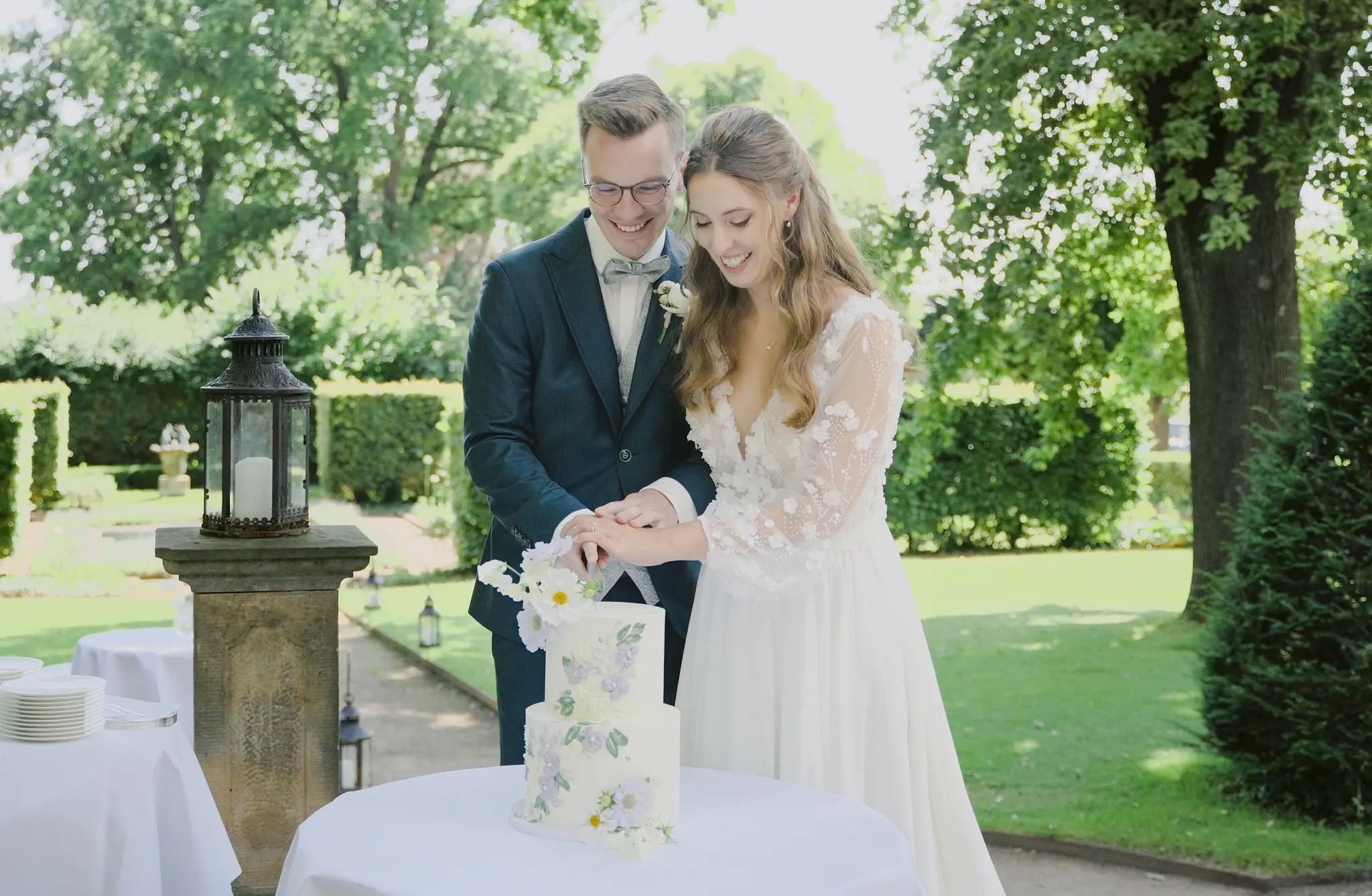 Das Brautpaar schneidet gemeinsam eine zweistöckige weiße Torte mit echten Blumen an.