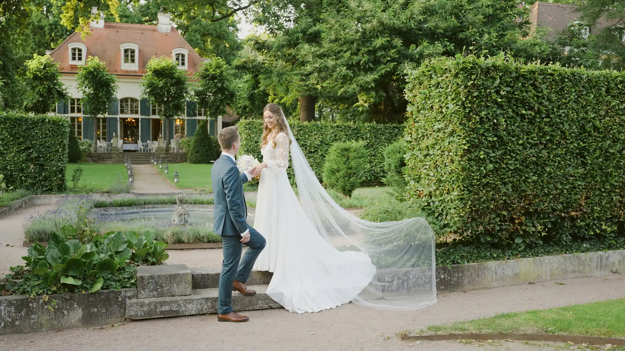 Weitwinkelaufnahme des Brautpaars auf einem Kiesweg im weitläufigen Garten der Villa.